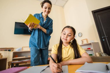 Mutlu öğretmen Montessori okulunda ders sırasında not defterine yazan Asyalı kıza bakıyor.