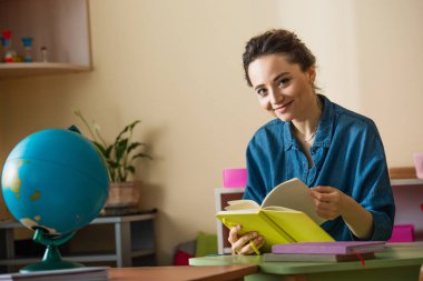 Montessori okulunda elinde kitapla dünyanın yakınındaki kameraya gülümseyen genç öğretmen.