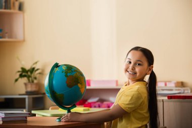 Montessori okulunda dünyanın yakınındaki kameraya gülümseyen neşeli Asyalı kız.