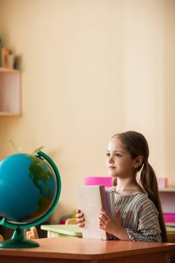 Montessori okulunda dünyanın yanında oturmuş kitap tutan bir kız.