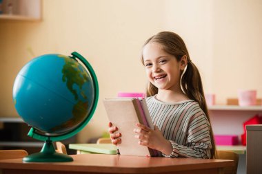 Montessori okulunda dünyanın yanında oturmuş kitap tutan mutlu bir kız.