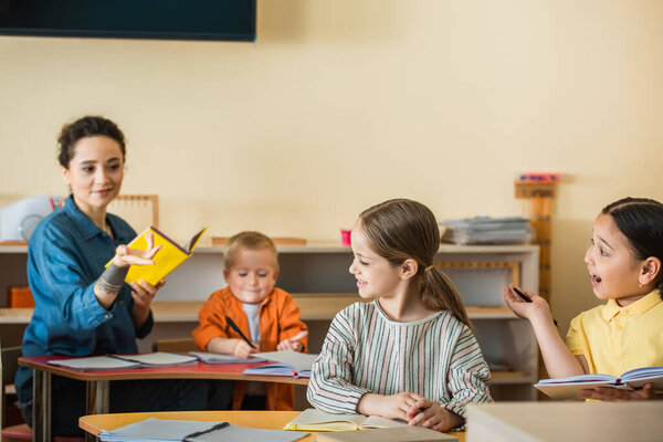 blurred teacher holding book and pointing with hand near interracial kids during lesson