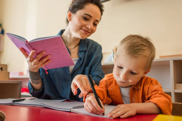 Profesor Sonriente Señalando Con Dedo Cerca Niño Escribiendo Dictado Escuela — Foto de Stock