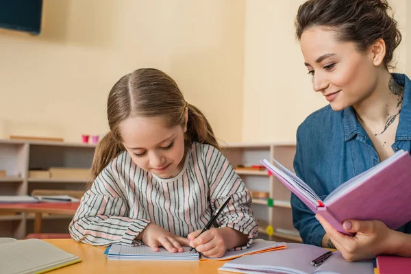 Glad Lärare Ler Nära Koncentrerad Flicka Skriva Diktamen Montessori Skolan — Stockfoto