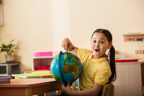amazed asian girl pointing at globe and looking at camera in montessori school