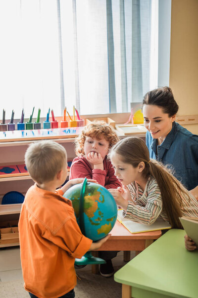 amazed girl looking at globe near boys and smiling teacher in classroom
