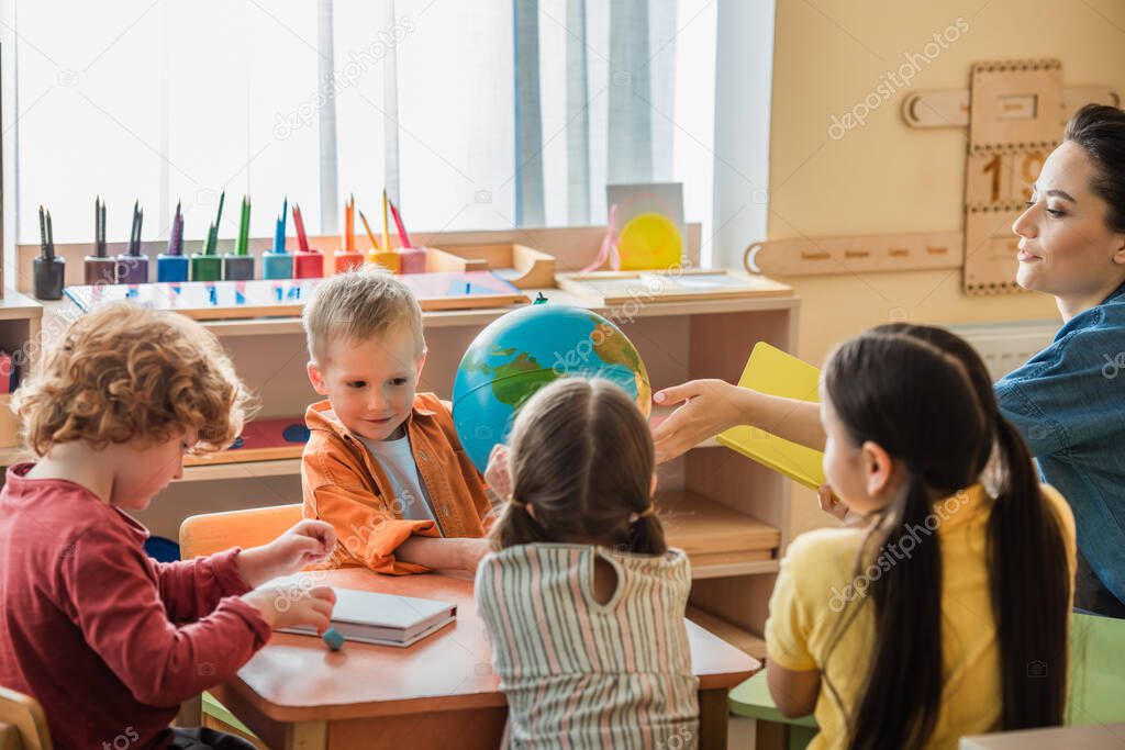 teacher pointing at globe while talking to kids during lesson in montessori school