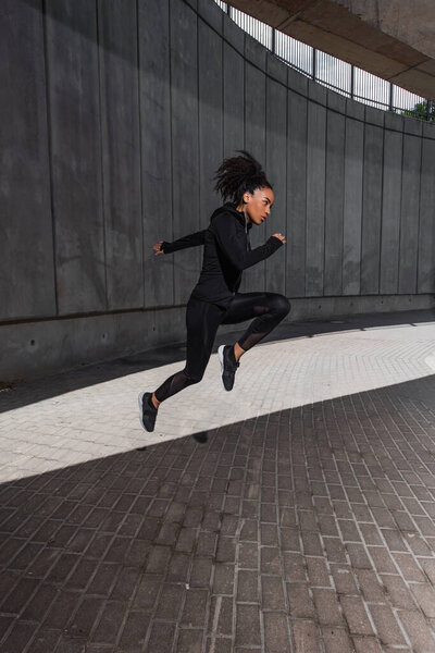 Side view of african american runner jumping near building on urban street 