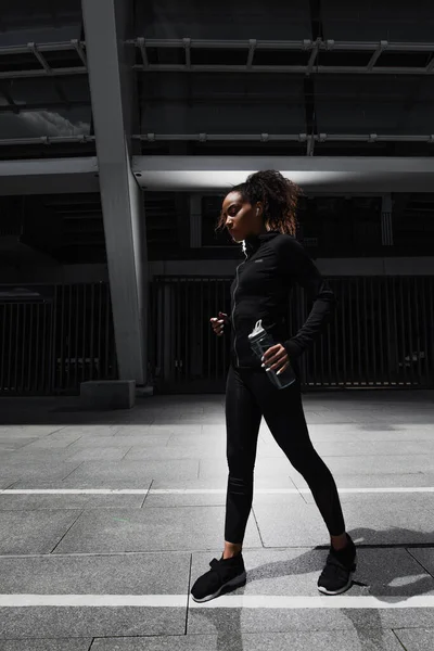 非洲裔美国女运动员的侧面照片 傍晚时分 她戴着耳机 在城市街道上拿着运动瓶 — 图库照片