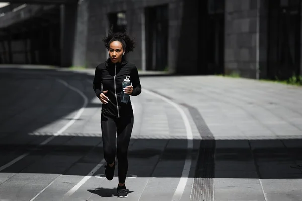 非洲裔美国人跑步者 带着装有耳机的运动瓶子在城市街道上听音乐 — 图库照片