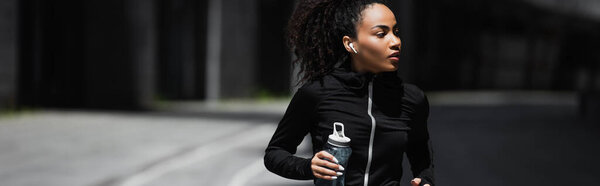 Young african american sportswoman in earphone holding sports bottle outdoors, banner 