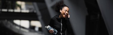 Cheerful african american woman in sportswear listening music in earphone and holding sports bottle outdoors, banner  clipart