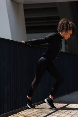 Side view of fit african american sportswoman in earphone warming up near bridge  clipart