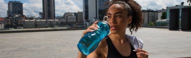 Afro-Amerikan sporcu kadın su içiyor ve şehir caddesinde havluyu tutuyor. 