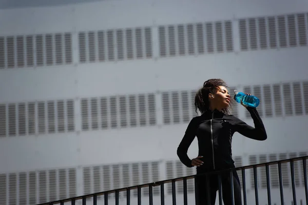 都市部の路上で水を飲むイヤホンでアフリカ系アメリカ人スポーツ女性の低角度ビュー — ストック写真