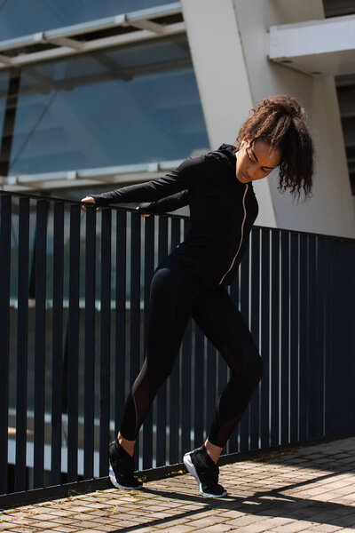 Fit african american sportswoman in earphone warming up beside bridge outdoors 