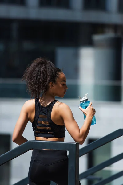 都市部の路上で水とスポーツボトルを保持するかなりアフリカ系アメリカ人のスポーツ女性の側面図 — ストック写真
