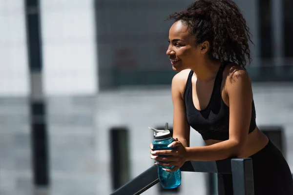 屋外の手すりの近くに笑みを浮かべてスポーツボトルを持つかなりアフリカ系アメリカ人のスポーツ女性 — ストック写真
