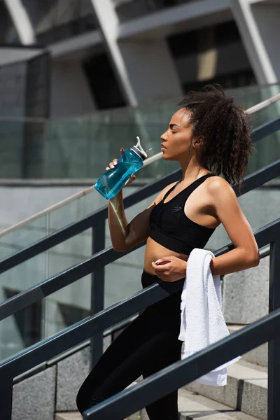 手すりの近くに水のボトルを保持タオルとかなりアフリカ系アメリカ人のスポーツ女性の側面図 — ストック写真