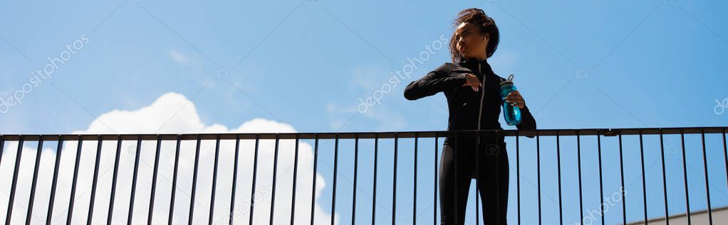 Low angle view of african american sportswoman in earphone and sportswear holding sports bottle on bridge, banner 