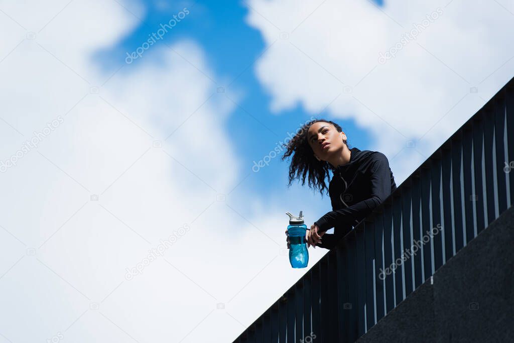 Low angle view of african american sportswoman holding sports bottle with sky at background 