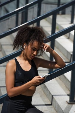 Kulaklıklı Afrikalı Amerikalı kadın merdivenlerde cep telefonu kullanıyor. 