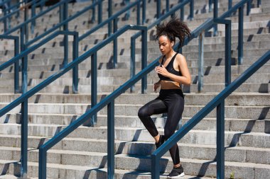 African american sportswoman in earphone running on stairs near railing on urban street  clipart