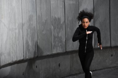 Odaklanmış siyah spor ceketli Afro-Amerikan kadın açık havada koşuyor. 