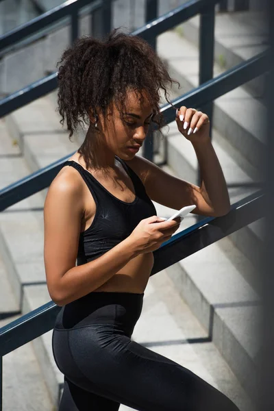 手すり近くのスマートフォンを使用して無線イヤフォンのアフリカ系アメリカ人スポーツ女性 — ストック写真