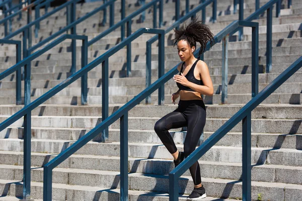 戴耳机的非洲裔美国女运动员在城市街道栏杆附近的楼梯上跑步 — 图库照片