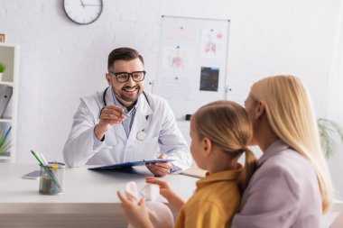 Gülümseyen çocuk doktoru elinde kalem ve not panosuyla anne ve çocuğun yanında. 
