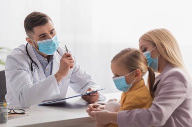 Tıbbi maskeli çocuk doktoru klinikte anne ve kızının yanında pano tutuyor. 