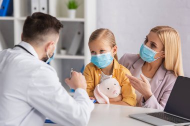 Tıbbi maskeli anne, bulanık doktor ve dizüstü bilgisayarın yanındaki kızı işaret ediyor. 