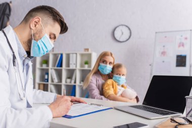 Tıbbi maskeli doktor, klinikteki aile ve cihazların yanında panoya yazı yazıyor. 