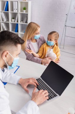Tıbbi maskeli çocuk dizüstü bilgisayar kullanarak anne ve aile doktorunun yanında konuşuyor.