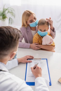 Koruyucu maskeli bir anne, aile doktorunun yanında reçete yazan bir oyuncakla çocuğa dokunuyor.
