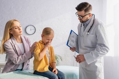Hasta çocuk elinde panoyla anne ve aile doktorunun yanında hapşırıyor. 