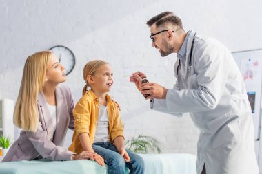 Doktor ağzı açık bir çocuğun yanında şurup tutuyor ve annesi klinikte. 
