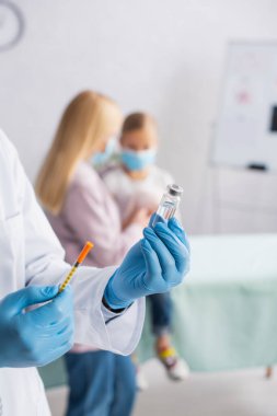 Elinde aşı ve şırınga olan çocuk doktoru anne ve çocuğu bulanıklaştırıyor. 