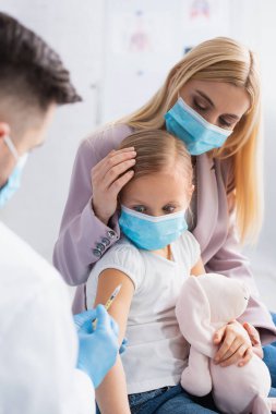 Tıbbi maskeli anne ve kızın yanında şırınga tutan bulanık çocuk doktoru. 