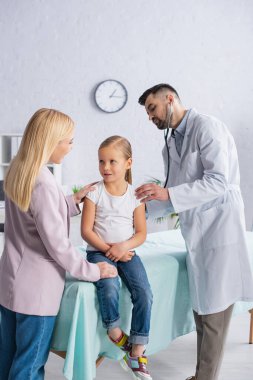 Doktor hastanede steteskop kullanırken annesi çocuğa sarılıyor. 