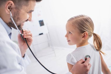Hastanede steteskopla çocuk doktoruna bakan gülümseyen bir çocuk. 