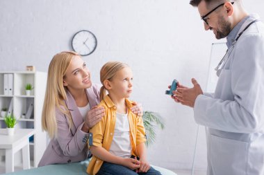 Gülümseyen anne, klinikte astım spreyiyle aile doktorunun yanında çocuğa sarılıyor. 