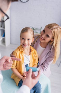 Bulanık aile doktoru, neşeli anne ve kızın yanındaki astım spreyini işaret ediyor. 