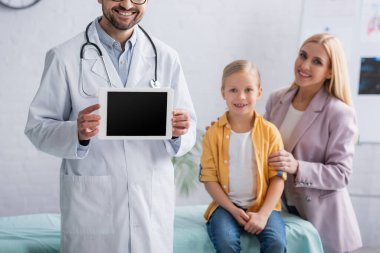 Gülümseyen aile doktoru arka planda anne ve çocuğun yanında dijital tablet tutuyor. 