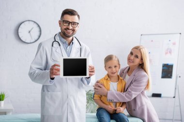 Annesinin yanında dijital tablet tutan pozitif aile doktoru tıbbi kanepede kızı kucaklıyor. 