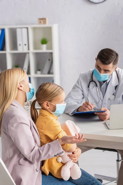 Woman hugging daughter and talking to doctor in medical mask writing on clipboard