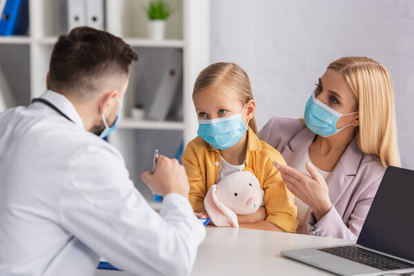 Мать в медицинской маске указывает на дочь рядом с размытым доктором и ноутбуком 