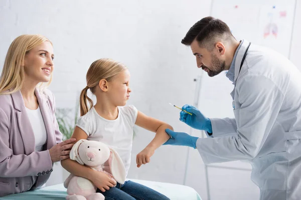 Sonriente Madre Pie Cerca Hija Haciendo Muecas Médico Con Jeringa —  Fotos de Stock