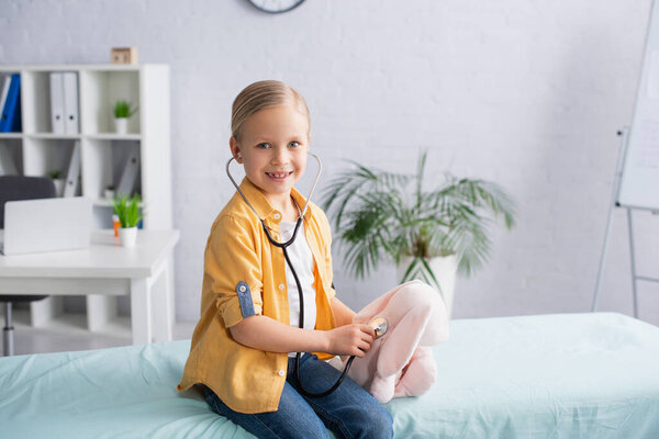 Улыбающийся ребенок в стетоскопе держит мягкую игрушку на медицинском диване 